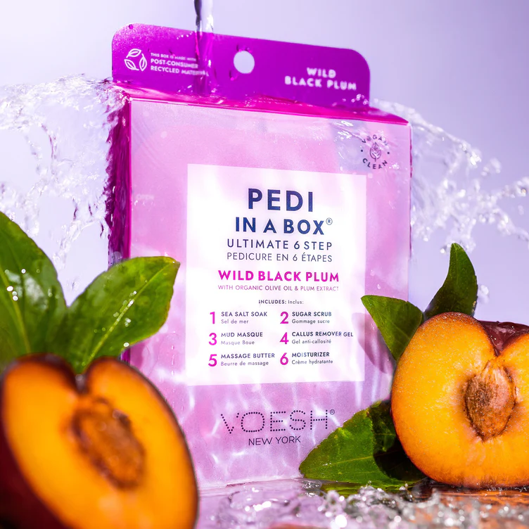 A pink package of "Pedi in a Box" ultimate 6-step pedicure in Wild Black Plum scent from VOESH New York is surrounded by fresh plums and splashed with water.