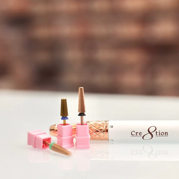 A close-up of four Cre8tion 5 in 1 Long Bit (3/32") with pink holders and a white nail drill handle also labeled "Cre8tion" resting on a white surface. The background is blurred.