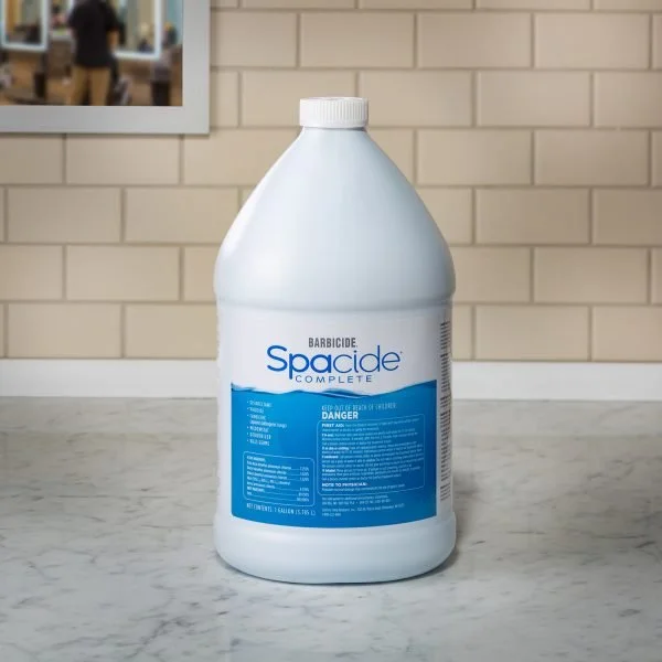 A large white plastic bottle of Barbicide Spacide Complete 1 Gallon on a marble countertop against a tiled wall background.