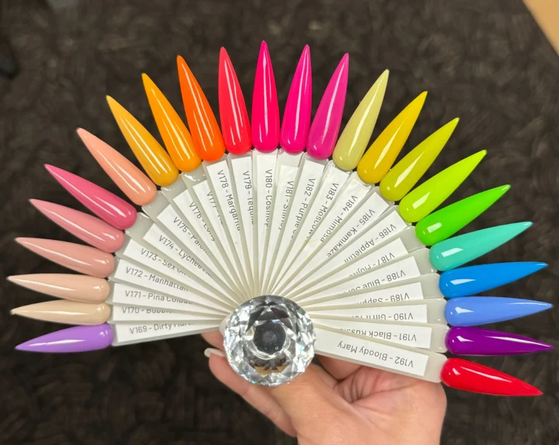 A hand holds a fanned-out set of artificial nails from the Nitro VIBE Collection 3-in-1 Powder, Gel, & Lacquer (24 Colors), showcasing 24 colors in vibrant hues. Each nail is labeled with its corresponding color code and name, featuring a clear decorative gem at the center, highlighting the exquisite Powder Gel Lacquer finish.
