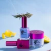 Three bottles of blue and pink nail lacquer and Nitro VIBE Collection 3-in-1 Powder, Gel, & Lacquer (24 Colors) with a rosemary sprig. A lemon slice and curl, and a rose gold cuticle pusher are also in the background on a light blue surface.