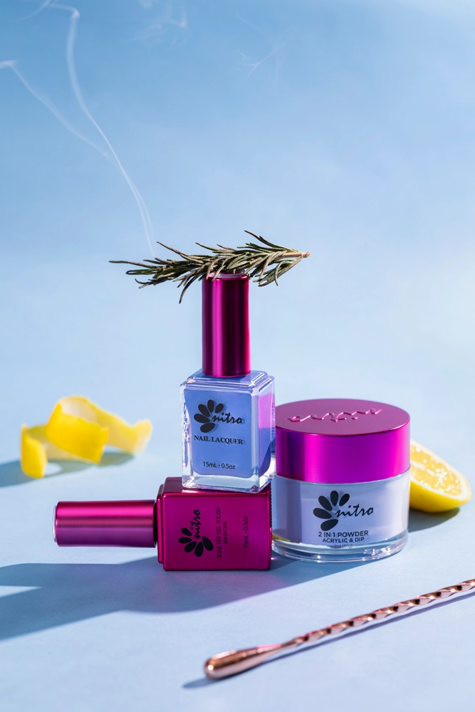 Three bottles of blue and pink nail lacquer and Nitro VIBE Collection 3-in-1 Powder, Gel, & Lacquer (24 Colors) with a rosemary sprig. A lemon slice and curl, and a rose gold cuticle pusher are also in the background on a light blue surface.