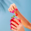 Two hands with bright pink Nitro VIBE Collection 3-in-1 Powder, Gel, & Lacquer (24 Colors) nails hold a glass of red iced beverage garnished with mint and a cherry against a blue background.