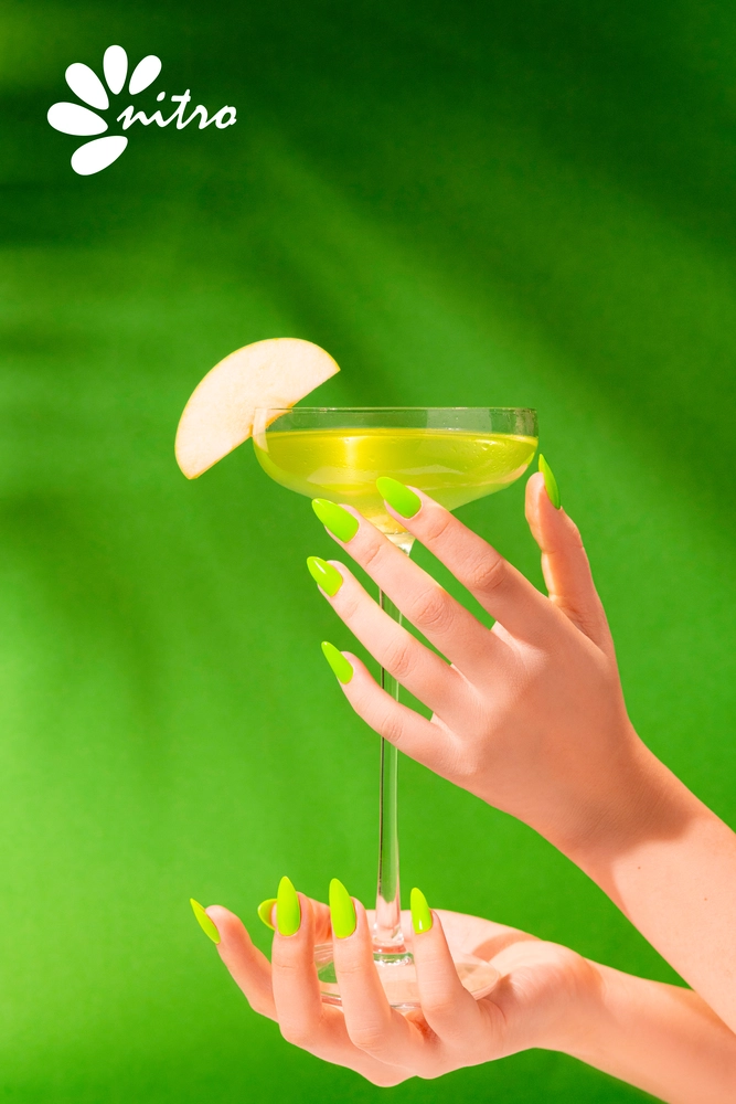 A hand with neon green nails, courtesy of the Nitro VIBE Collection 3-in-1 Powder, Gel, & Lacquer (24 Colors), holds a cocktail glass garnished with a slice of apple against a bright green background. The "Nitro" logo is visible in the top left corner.