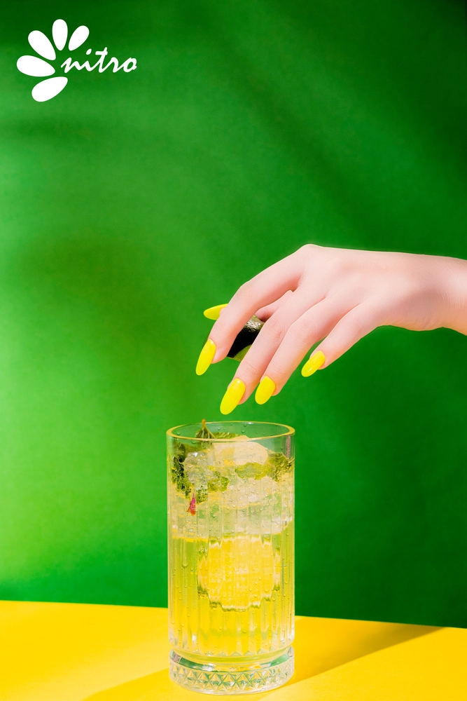 A hand with yellow painted nails from the Nitro VIBE Collection 3-in-1 Powder, Gel, & Lacquer (24 Colors) squeezes a lime slice over a glass of ice-filled mojito against a green background. "Nitro" text and logo are visible in the top left corner. The vibrant 24 colors offer endless options, including this striking Powder Gel Lacquer.