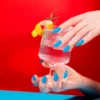 A hand with blue painted nails, from the Nitro VIBE Collection 3-in-1 Powder, Gel, & Lacquer (24 Colors), holds a cocktail garnished with a lemon twist and rosemary in a glass against a red background, with the "nitro" logo in the top left corner.