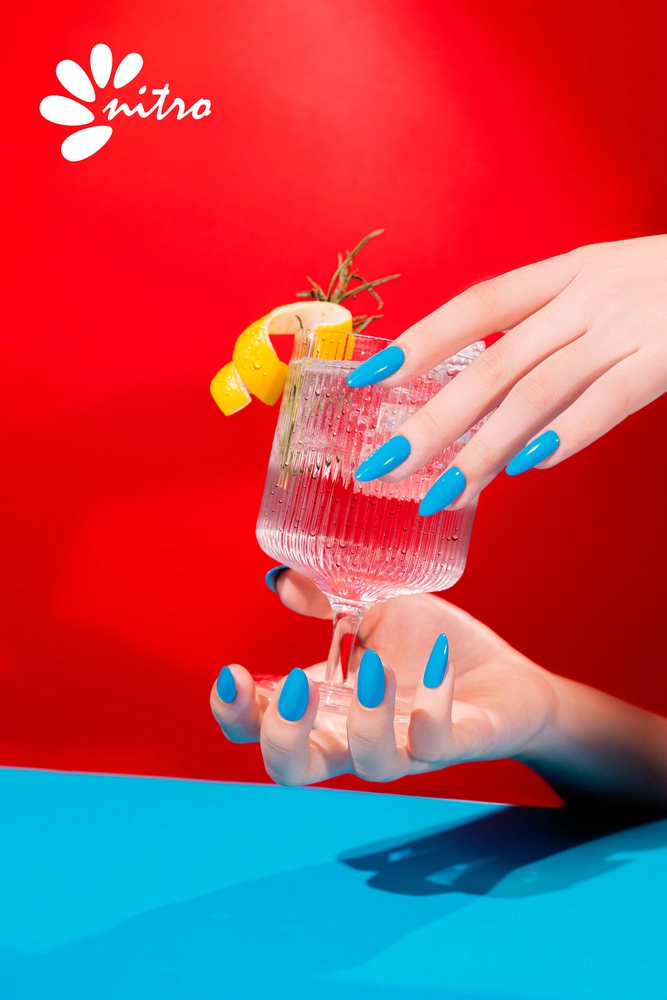 A hand with blue painted nails, from the Nitro VIBE Collection 3-in-1 Powder, Gel, & Lacquer (24 Colors), holds a cocktail garnished with a lemon twist and rosemary in a glass against a red background, with the "nitro" logo in the top left corner.