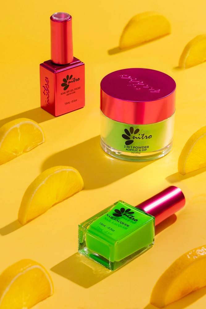 Brightly colored Nitro VIBE Collection 3-in-1 Powder, Gel, & Lacquer (24 Colors) bottles and a jar on a yellow background, surrounded by lemon wedges. One Nitro VIBE bottle and jar are pink with green accents; the other bottle is green with a pink cap.