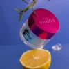 A jar with a pink lid labeled "Nitro VIBE Collection 3-in-1 Powder, Gel, & Lacquer (24 Colors)" is hovering above a halved lemon against a blue background, with ice cubes and a sprig of greenery in the scene.