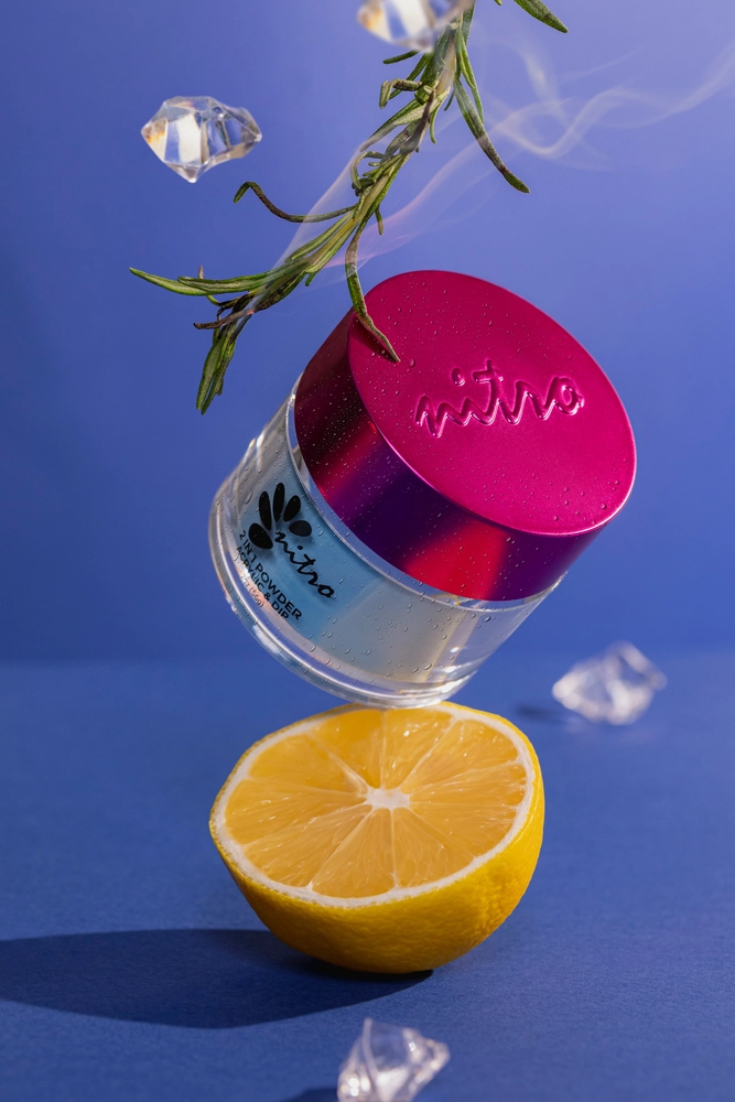 A jar with a pink lid labeled "Nitro VIBE Collection 3-in-1 Powder, Gel, & Lacquer (24 Colors)" is hovering above a halved lemon against a blue background, with ice cubes and a sprig of greenery in the scene.