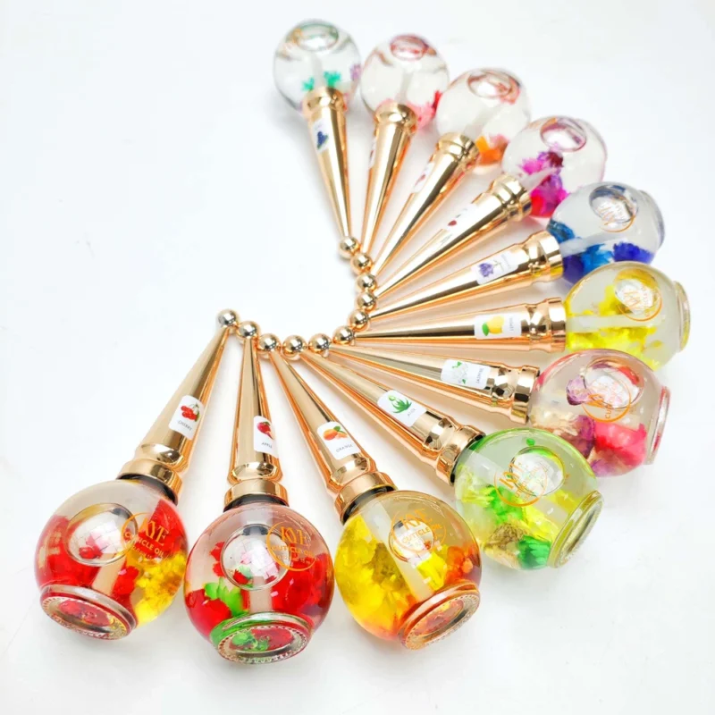 A set of twelve ballpoint pens arranged in a circle. Each pen has a clear, round top filled with colorful decorations and a gold-colored body, reminiscent of the elegant design seen in KYE Flower Cuticle Oil 0.9oz packaging.