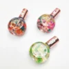 Three spherical bottles of KYE Cuticle Oil 0.9oz with copper-colored caps, each containing different colored floral designs, sit elegantly on a white surface. These 0.9oz bottles add a touch of beauty to your manicure routine.