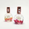 Two bottles of KYE Flower Cuticle Oil 0.9oz with gold caps. One, labeled "Peach" with yellow flowers inside, and the other, labeled "Strawberry" with pink flowers inside. Each 0.9oz bottle nourishes your cuticles while adding a touch of floral elegance.