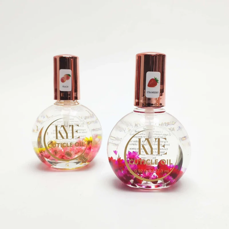 Two bottles of KYE Flower Cuticle Oil 0.9oz with gold caps. One, labeled "Peach" with yellow flowers inside, and the other, labeled "Strawberry" with pink flowers inside. Each 0.9oz bottle nourishes your cuticles while adding a touch of floral elegance.