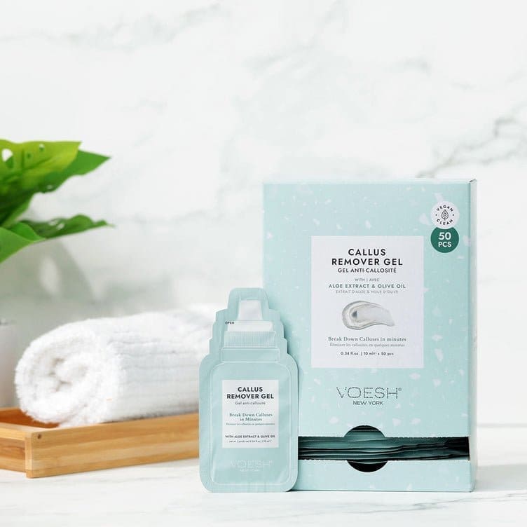 A product display featuring Voesh Callus Remover Gel Aloe Extract & Olive Oil Single Pack next to a rolled white towel on a wooden tray, against a clean, white background with a green plant.