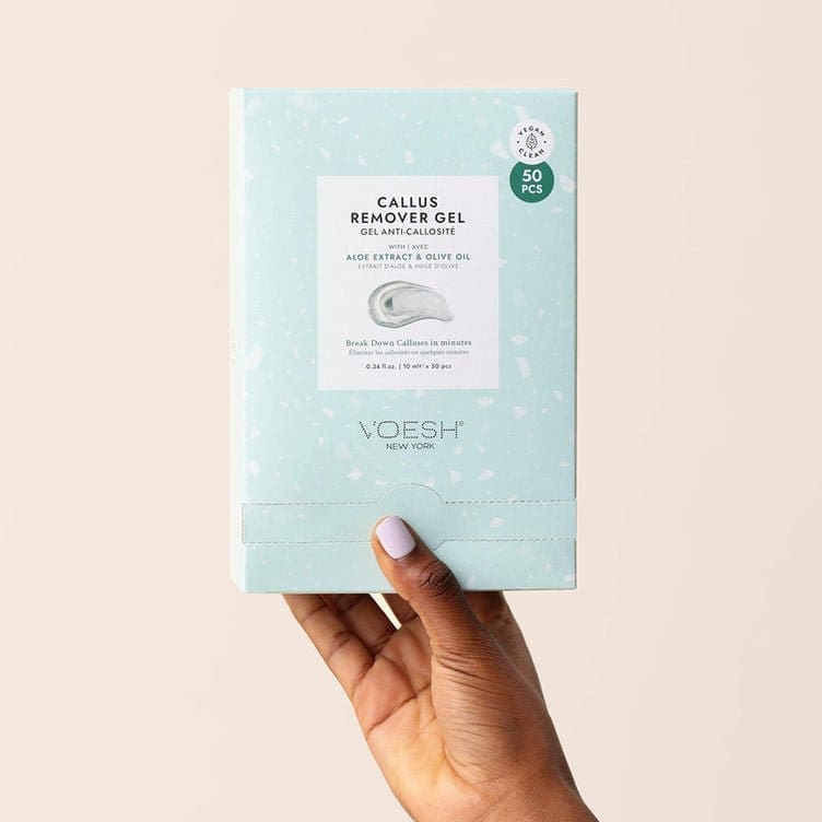 A hand holding a box of Voesh Callus Remover Gel Aloe Extract & Olive Oil Single Pack, containing 50 pieces. The light blue box boasts white speckled design elements.