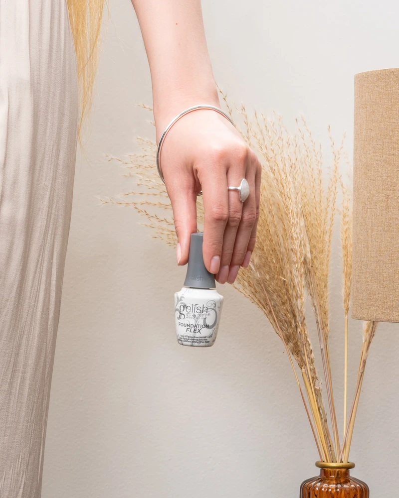 A hand holding a bottle of Gelish Soak-Off Gel Polish near a decorative vase with dried grasses, complemented by the reliable Gelish Foundation Flex Soak-Off Rubber Base Nail Gel - Clear 15ML for perfect nails.