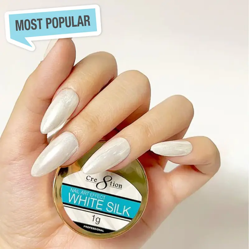 A hand with white, oval-shaped nails carefully cradles a small round container labeled "Cre8tion White Silk Chrome Nail Art Effect 1g." A speech bubble in the top left corner announces "Most Popular." Experience the luxurious shine of the White Silk Chrome effect for your nail art.